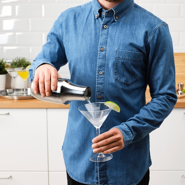 Person pouring drink from stainless steel shaker into cocktail glass with lime wedge.