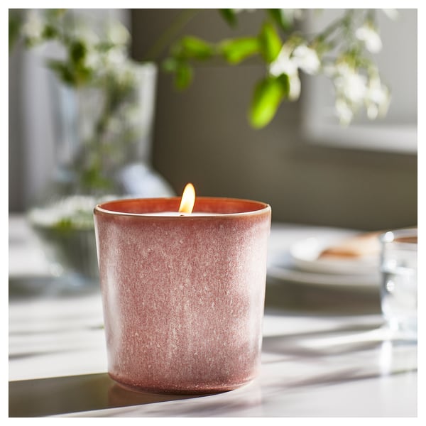 Lit LUGNARE pinkish ceramic candle, textured finish, glowing flame, on table with blurry blurred greenery and plate.