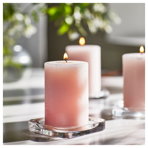 Three lit pink candles on clear glass holders, set on a white surface, surrounded by blurred greenery.