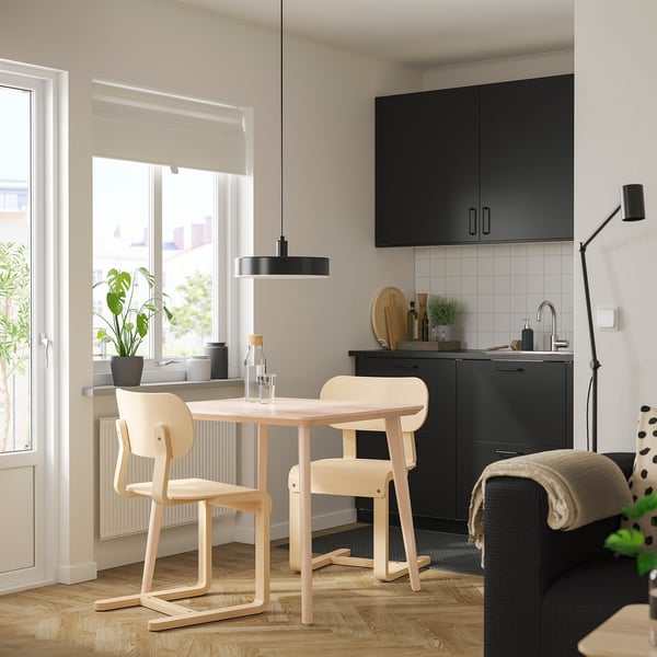 Modern kitchen with black cabinets, wooden table, & light chairs by window, cozy and well-lit.