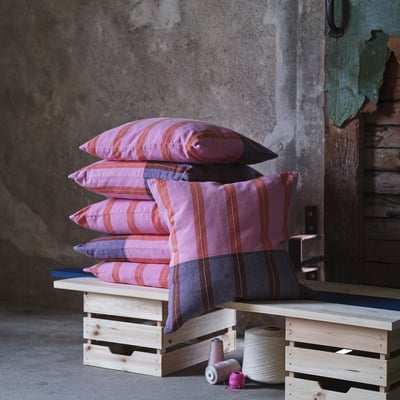 Stack of pink and orange striped MÄVINN cushions on wooden crate, spools below.