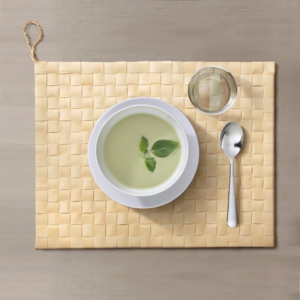 A bowl of green soup on a handwoven, rectangular palmyra leaf place mat with a spoon and small dish. The mat has a loop for hanging.