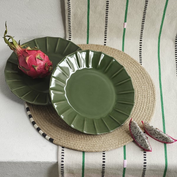 Green MÄVINN stoneware plate sits on round woven placemat with dragon fruit. Plates scalloped edge adds texture, slightly larger than fruit.