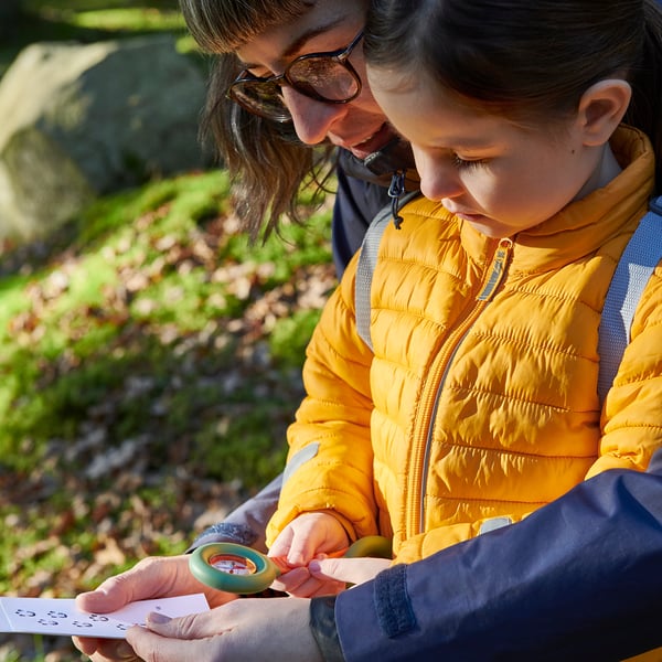 A child and an adult examine a compass with a magnifying glass outdoors.