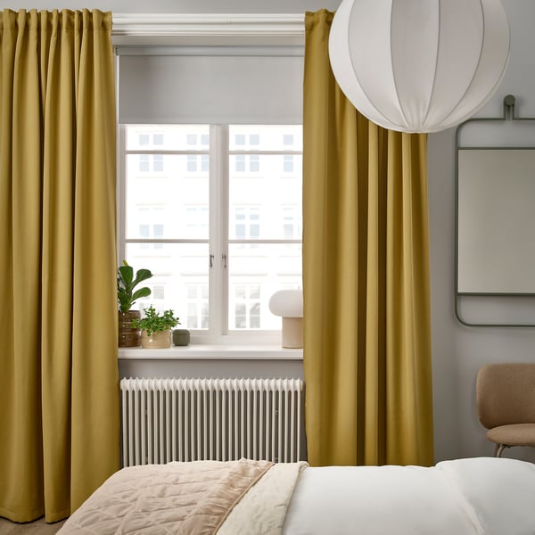 Modern bedroom with MAJGULL mustard curtains, white bed, cosy chair, and large lamp. Curtains tower over window and bed.
