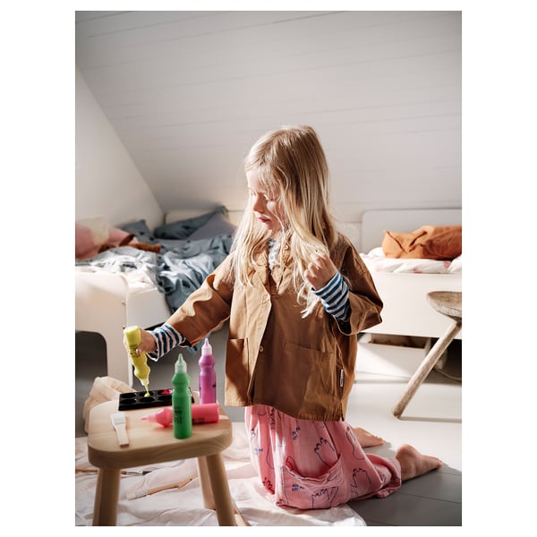 A young girl is crafting with the MÅLA art set featuring brushes, scissors, and markers on a wooden stool in a room with beds.
