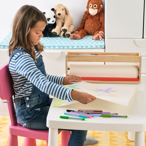 A girl sits at a kids desk, drawing on a paper roll. The holder organises pens and crayons. Stuffed animals are visible on a shelf behind the person.