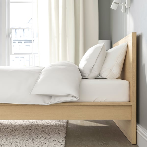 A wooden bed frame with a high headboard and white bedding against a curtained window, providing a comfortable backrest for sitting and reading.