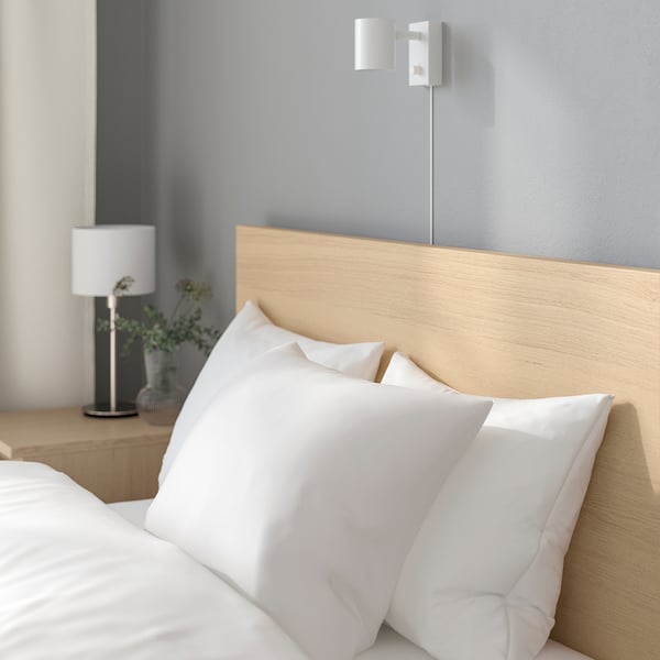 A modern bed with a wooden headboard, white pillows, and a bedside lamp. The lamp provides adjustable lighting for reading.