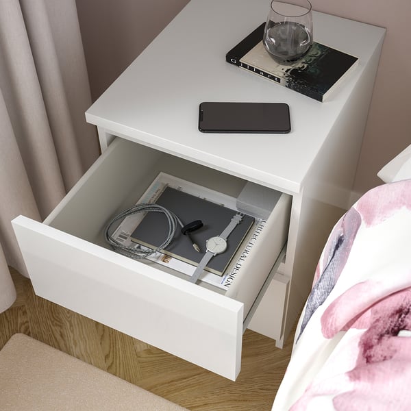 White bedside table with open drawer showing watch, charging cable, and books; smartphone and wine glass on top.