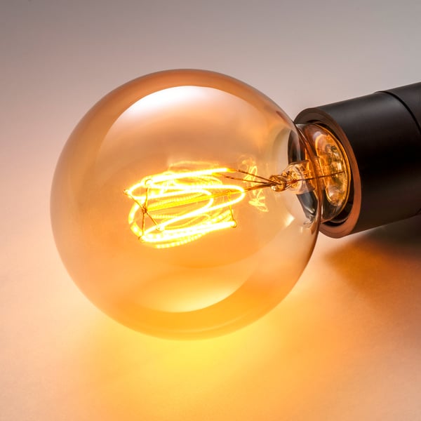 Illuminated vintage-style bulb in amber glass, glowing warmly with intricate internal filaments, attached to a black base.