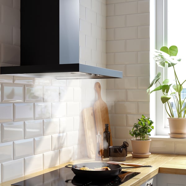 Modern kitchen with black hood, white tiles, wooden countertop, potted plants, and a cooking pan.