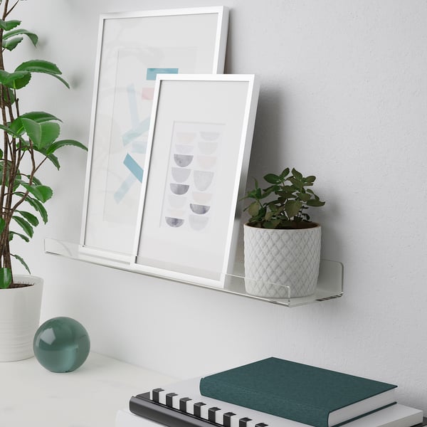 Clear wall shelf holds picture frames and plants. Frames lean against wall, showcasing art.