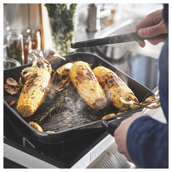Person grilling corn in black MIDDAGSMAT pan on a white stove. Corn is golden brown with charred husks.