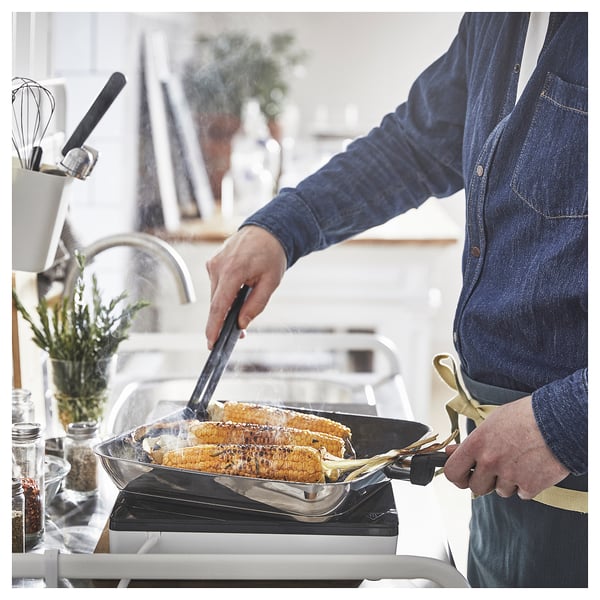 Person grilling corn in a kitchen, using a MIDDAGSMAT pan with a non-stick coating. The individual wears a blue apron, focusing on cooking.