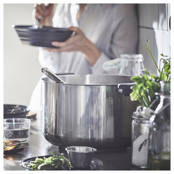 Person holding plates near a large, shiny silver stockpot on a stove, surrounded by cooking ingredients and utensils.