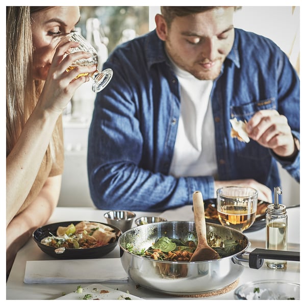 A couple eats meal near a sauté pan on a table. Person drinks from a glass, person eats with a fork.