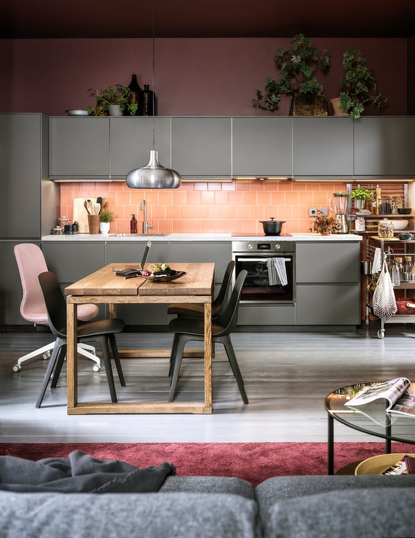 Modern kitchen with wooden table, grey chairs, pendant light, plants, and grey cabinets.