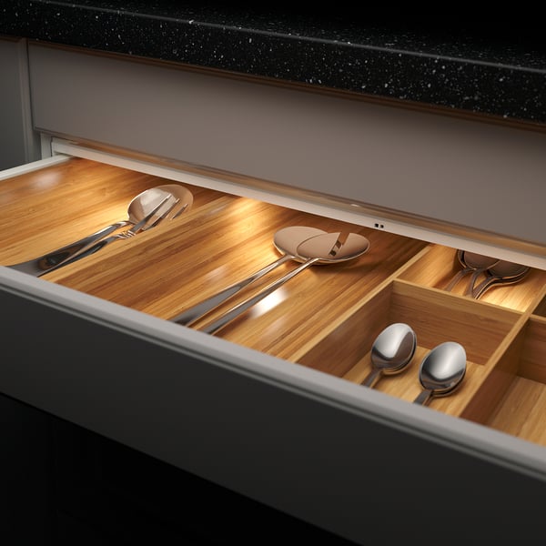 Open kitchen drawer with wooden compartments, holding various metallic utensils. Built-in warm led lighting illuminates contents.
