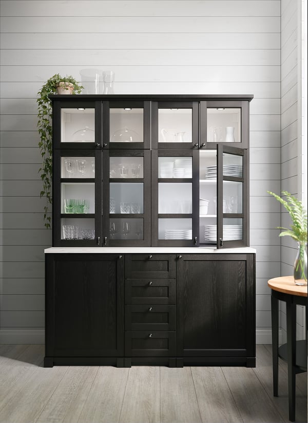Modern black kitchen cabinet with glass doors, led lights, housing various dinnerware, and a wooden countertop.
