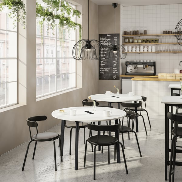 Modern café with round white tables, black chairs, large windows, and hanging lights. Tables have coffee cups and menu board behind.