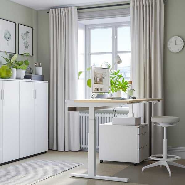 Home office setup with MITTZON white sit/stand desk by large window, white cabinet, clock, plants, and chair.