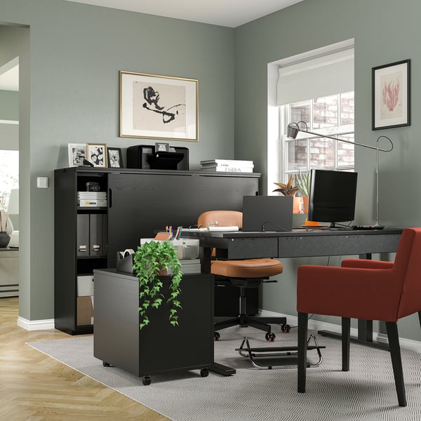 Modern home office with black MITTZON desk, red chair, green walls, plants, and artwork.
