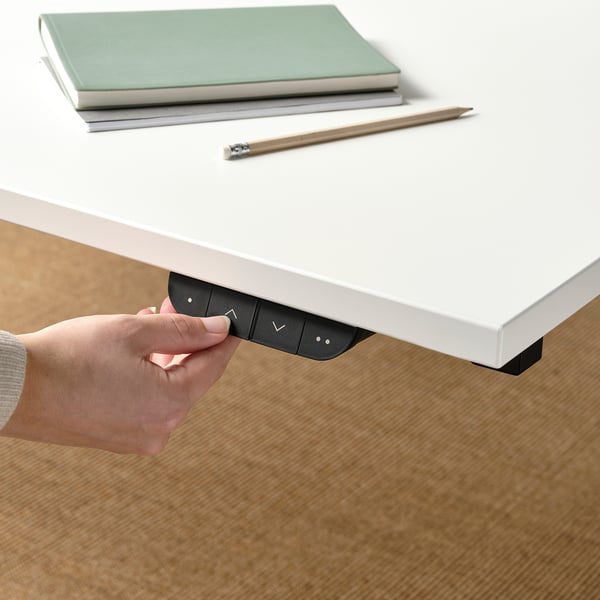 Hand adjusting electric sit/stand desk controls; laptop and pen on top.