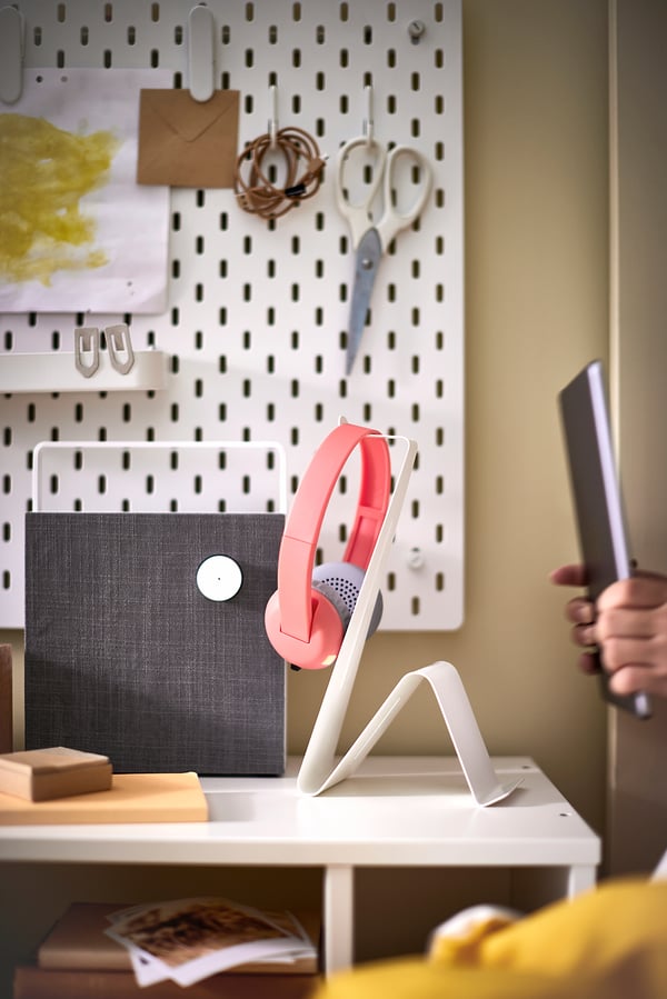 A desk with a white stand holding pink headphones and a tablet. A pegboard with office supplies in the background.