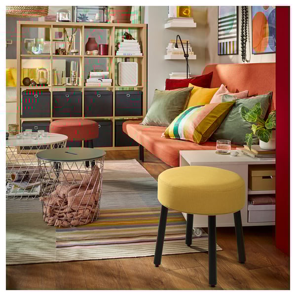 Vibrant living room with orange sofa, colourful pillows, yellow stool, wire table, striped rug, and decorated shelves.