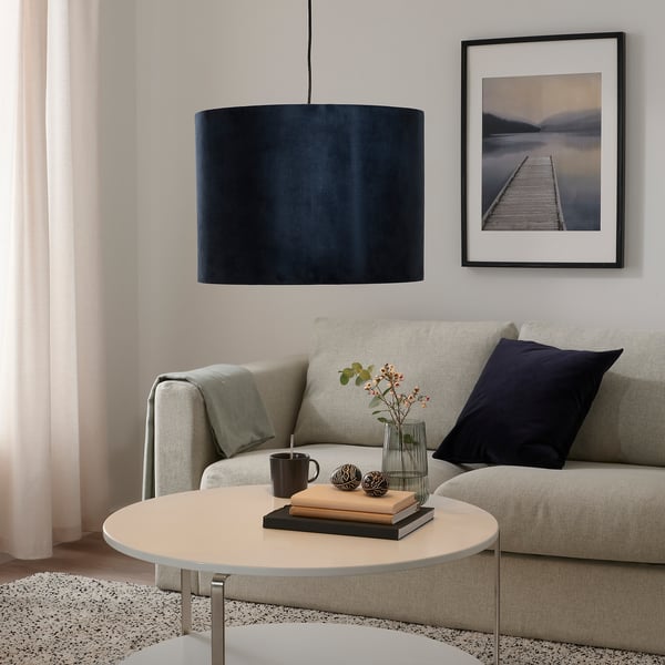 Modern living room with large blue velvet lampshade over grey sofa, warmly lit. Coffee table with books, mug, and flowers. Wall has dock picture.