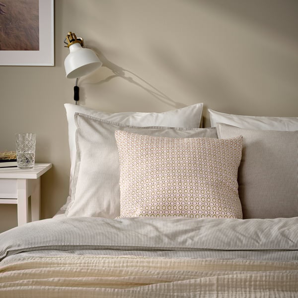 A MOROTSMOTT pillow on a bed. The pillow is light pink with a woven pattern. Its smaller than the bed and surrounded by larger white pillows.