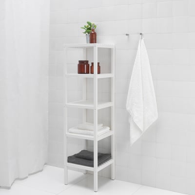 A tall white bathroom shelf unit holds towels, bottles, and a plant, optimising space with multiple open shelves.