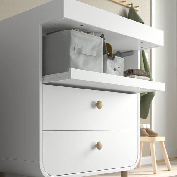 A white changing table with drawers and shelves. A practical storage shelf above the drawers, holding boxes and a cloth bag.