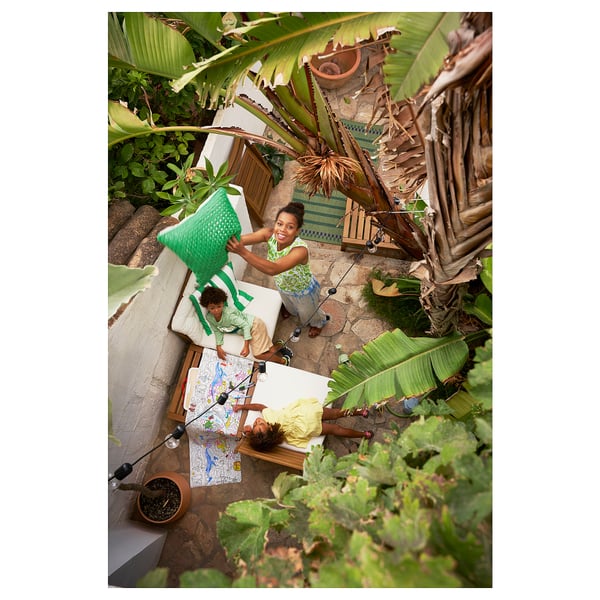 Three children play in an outdoor space with a green NÄMMARÖ chair, surrounded by lush tropical plants.