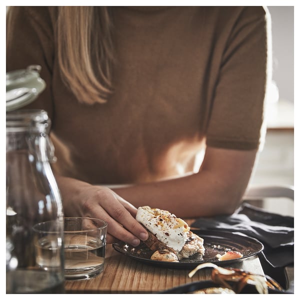 A person in a brown shirt holds a plate with food. On the table, theres a clear glass and a green bottle. The plate and glass have circular patterns.