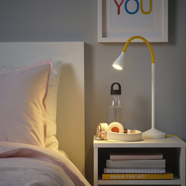 Bedroom with bed, yellow NÄVLINGE lamp, water bottle, and books.