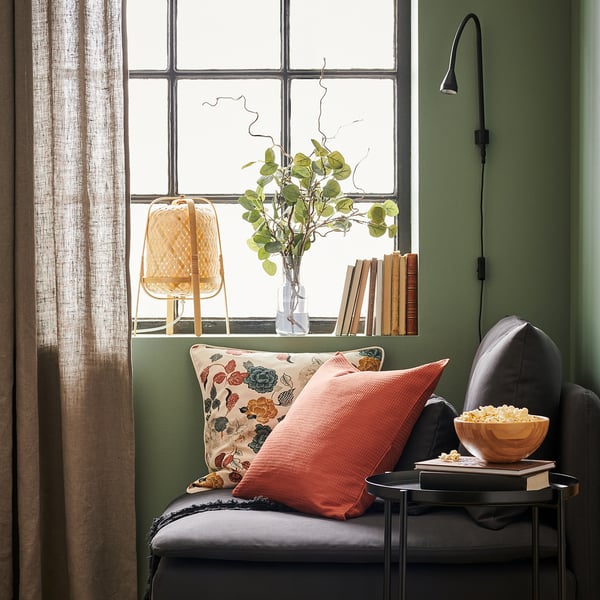 A cosy chair by a green window with an orange pillow, bowl of popcorn, lamp, and plant.