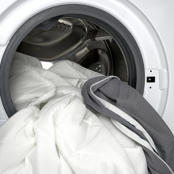 A white washing machine with a grey and white mattress pad inside, showcasing its machine-washable cover.