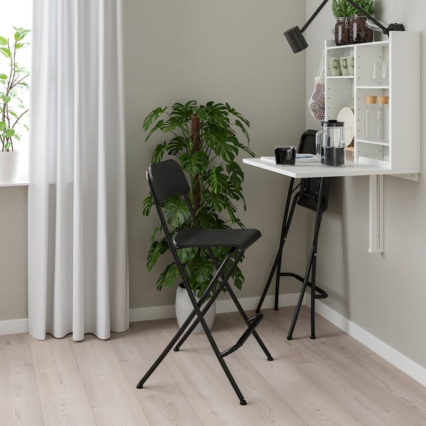 Minimalist home office: white wall-mounted desk, black folding chair. Greenery, white curtains. Compact, efficient.