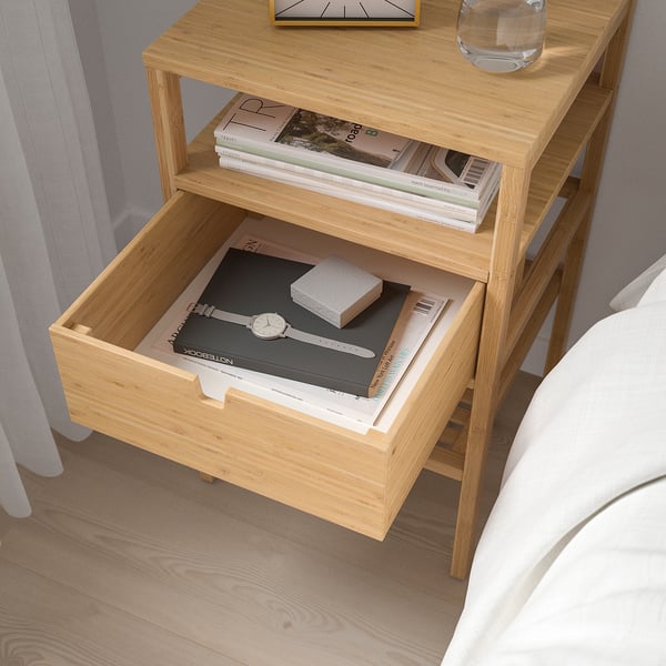 Wooden bedside table with open drawer showing books, glass, and clock on top. Drawer has handles on both sides.