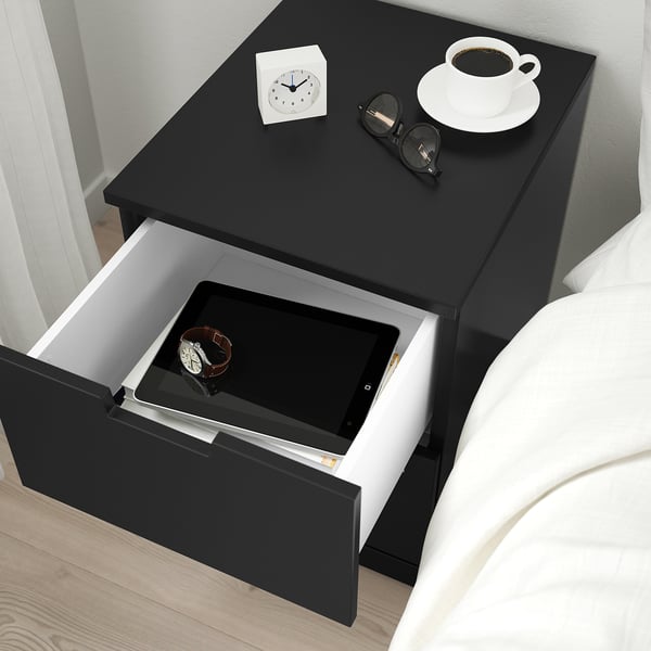 Black bedside table with open drawer showing tablet and watch near bed.
