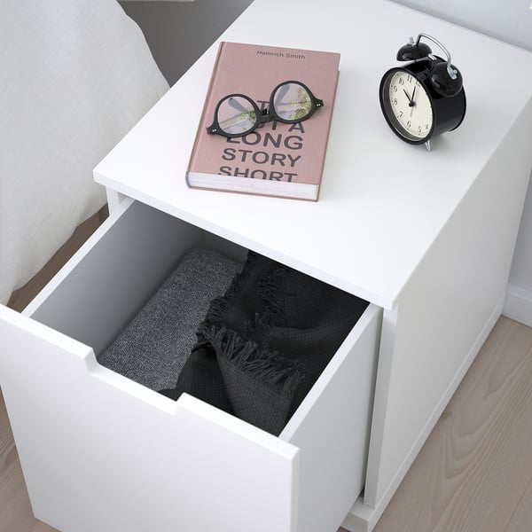 White bedside table with open drawer showing blanket, next to book and alarm clock.