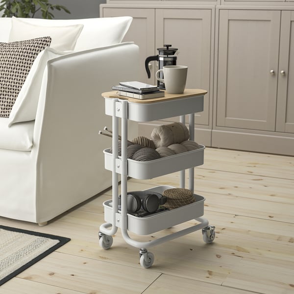 Small white rolling cart, perfect for storage, fits near sofa. Top has wooden lid, bottom has bins. Holds mugs, books, baskets.