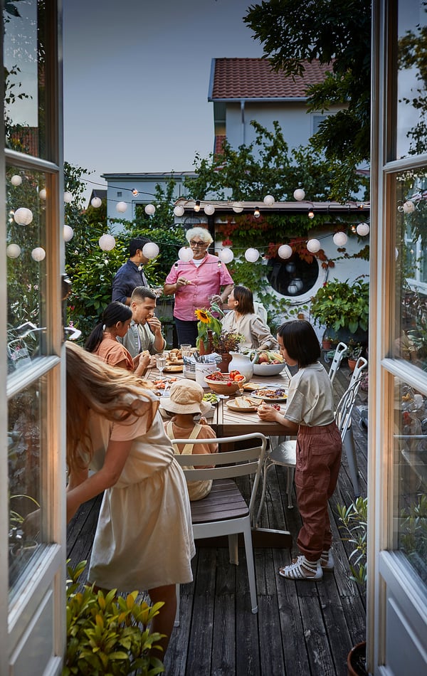 Family dines outdoors at long table. NORRMANSÖ chairs, pink and beige clothes, hanging lights, greenery.
