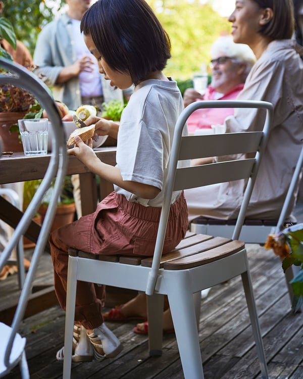 A young boy sits in a light grey metal and wood chair. The person eats a sandwich at an outdoor table surrounded by family.