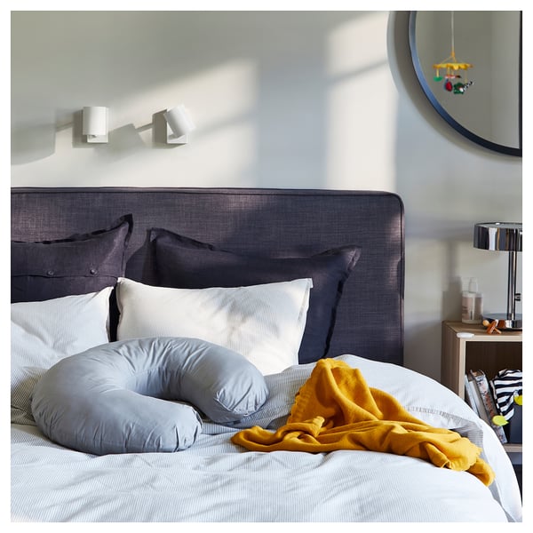 Modern bedroom with grey headboard, pillows, pregnancy pillow, yellow blanket, round mirror, and wooden nightstand with lamp.