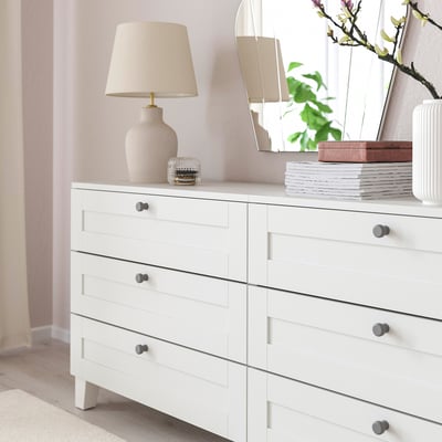 White dresser with six drawers and grey knobs, topped with a lamp, books, and a vase.