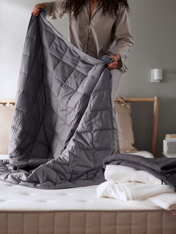 Person holding grey ODONVIDE weighted blanket over bed. Lightly padded with box-stitch pattern, recycled polyester and cotton.