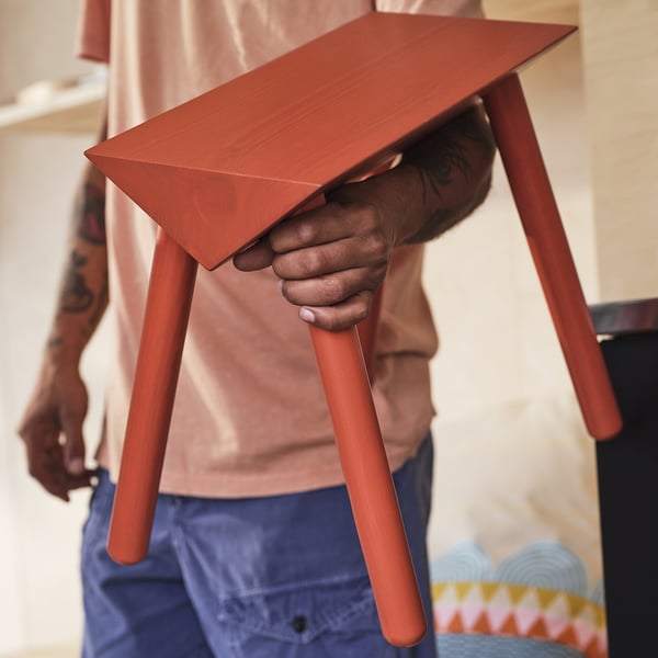Person holding a bright red, triangular-top, three-legged stool made of solid pine.